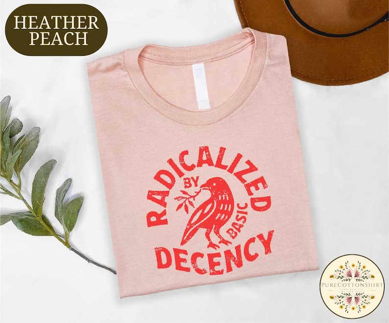 May include: A heather peach t-shirt with a crew neck, featuring a red graphic of a bird holding a branch, encircled by the words "RADICALIZED BY BASIC DECENCY." The shirt is folded and displayed on a white surface.