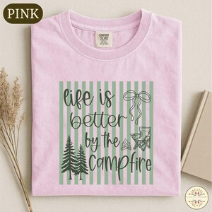 A pink t-shirt with a graphic design featuring a green and white striped pattern, pine trees, and camping-related elements, along with the text "life is better by the campfire".