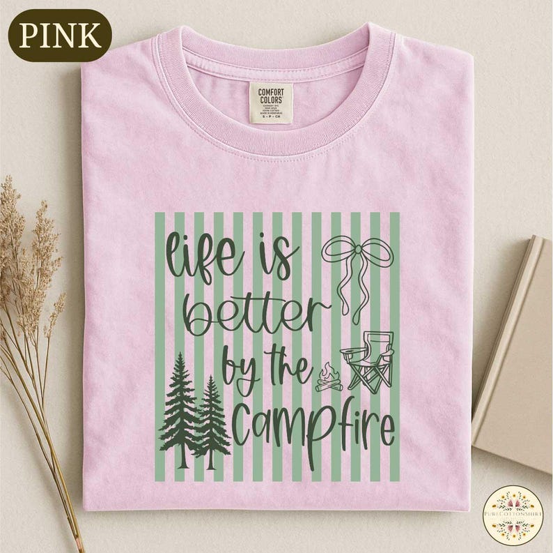 A pink t-shirt with a graphic design featuring a green and white striped pattern, pine trees, and camping-related elements, along with the text "life is better by the campfire".
