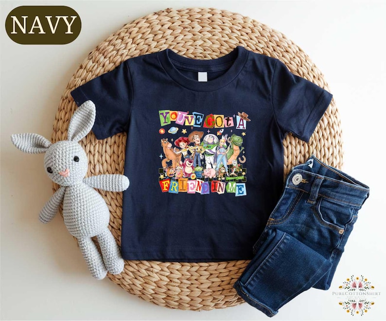 A navy blue t-shirt with a colorful graphic of characters from Toy Story, along with a crocheted gray rabbit toy and a pair of blue jeans, all arranged on a woven placemat.