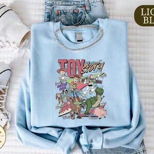 A light blue T-shirt with a cartoon character design, a necklace, and a pair of white sneakers are laid out on a bed.