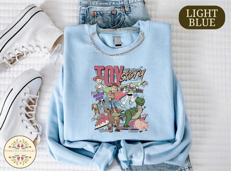 A light blue T-shirt with a cartoon character design, a necklace, and a pair of white sneakers are laid out on a bed.