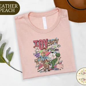 a pink t-shirt with a colorful graphic design of characters from the Toy Story movie, including Woody, Buzz Lightyear, and other beloved characters. The t-shirt is laid out on a white surface, with a brown hat and some greenery visible in the background.