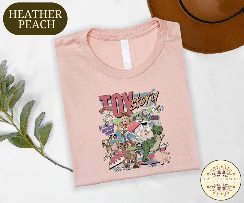 a pink t-shirt with a colorful graphic design of characters from the Toy Story movie, including Woody, Buzz Lightyear, and other beloved characters. The t-shirt is laid out on a white surface, with a brown hat and some greenery visible in the background.
