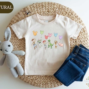 A white t-shirt with a Disney character design, including Mickey Mouse, Buzz Lightyear, Woody, and other popular characters, along with balloons, is displayed on a woven placemat. Next to the t-shirt is a crocheted stuffed rabbit, and a pair of blue jeans is also present.