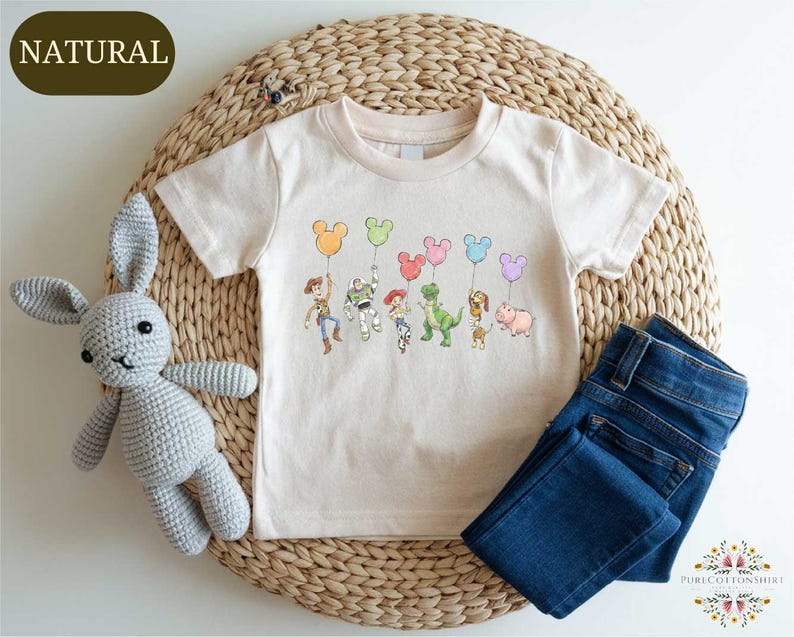 A white t-shirt with a Disney character design, including Mickey Mouse, Buzz Lightyear, Woody, and other popular characters, along with balloons, is displayed on a woven placemat. Next to the t-shirt is a crocheted stuffed rabbit, and a pair of blue jeans is also present.