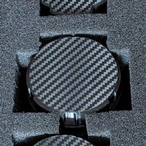 May include: Three round, carbon fiber patterned objects are nestled in a dark gray foam insert. The objects have a black and gray woven design. The foam insert has cutouts to hold each object securely.