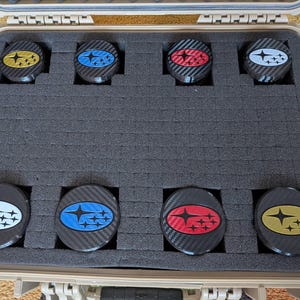 May include: A tan hard-shell carrying case with dark gray foam inserts containing eight circular emblems. Each emblem features a carbon fiber texture and the Subaru star logo in various colors including yellow, blue, red, white, and black.