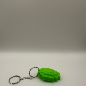 Peut inclure: Un porte-clés vert vif, pixélisé, avec un porte-clés et une chaîne argentés. Le porte-clés présente un design géométrique et anguleux. Le fond est uni, gris clair.