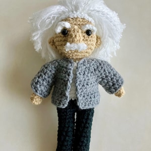 May include: A handmade crochet doll depicting Albert Einstein. The doll features a tan face with a white mustache and hair, wearing a gray jacket, white shirt, and dark green pants. The doll is approximately 8 inches tall.