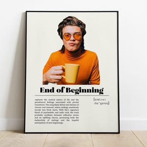 以下が含まれることがあります： 「End of Beginning」のテキストと、オレンジ色のサングラスとタートルネックを着て黄色のマグカップを持っている人の写真が特徴の額入りポスター。ポスターにはタイトルの下に説明文が含まれています。