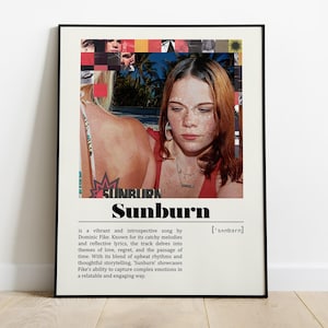 May include: A framed poster featuring a woman with red hair and freckles. The poster has the word "Sunburn" in large black letters, with a description below. The poster is in a black frame and is leaning against a white wall.