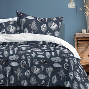 Peut inclure: Un ensemble de housse de couette et d'oreillers bleu marine avec un drap blanc. La literie présente un motif répété de divers coquillages dans des tons de blanc et de gris. La chambre a un mur bleu et une table de chevet en bois.