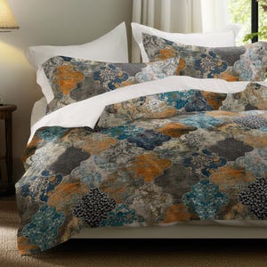 May include: A bed with a patterned comforter and pillowcases. The bedding features a mosaic design in shades of blue, orange, and gray. White sheets and pillowcases are visible. A framed picture hangs on the wall, and a lamp sits on a wooden side table.