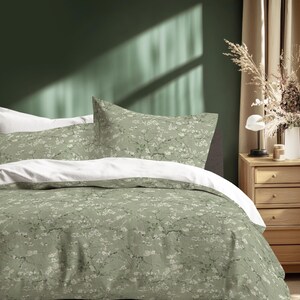 May include: A bed with a sage green floral patterned duvet cover and pillowcases. The bedding has a white underside. The room has a green wall and a wooden nightstand with a lamp and a vase of dried flowers.