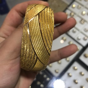 Op de afbeelding: Een brede, getextureerde gouden armband met een geweven ontwerp. De armband heeft een heldere, glanzende afwerking en wordt in een hand gehouden. De achtergrond toont andere sieraden.