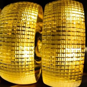 Könnte beinhalten: Zwei goldfarbene, breite, strukturierte Armreifen. Die Armbänder haben ein sich wiederholendes Muster aus kleinen, rechteckigen Blöcken, wodurch sie ein strukturiertes Aussehen erhalten. Die Armreifen werden vor einem dunklen Hintergrund präsentiert, der ihr glänzendes, metallisches Finish hervorhebt.
