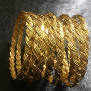May include: A collection of gold-colored bangles with a braided rope design. The bangles are stacked together, showcasing their intricate texture and reflective surface. The jewelry is displayed against a dark background.