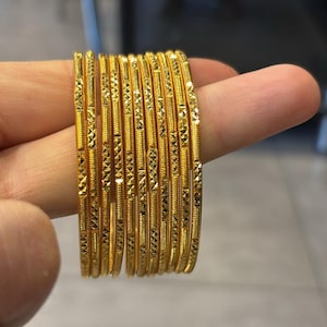May include: A collection of gold bangles, each with a detailed, textured design. The bangles are stacked together, showcasing their intricate patterns and shiny, reflective surface. The jewelry is held up by a finger.
