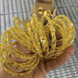 Peut inclure: Plusieurs bracelets dorés avec des motifs tressés et texturés complexes. Les bracelets sont tenus dans une main, avec une boîte en carton contenant d'autres bijoux en or en arrière-plan. Les bracelets ont une surface brillante et réfléchissante.
