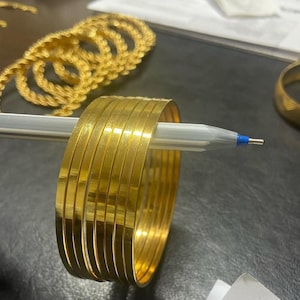 May include: A stack of gold-colored bangles, each with a flat, polished surface. The bangles are held together by a pen. Other gold jewelry is visible in the background.