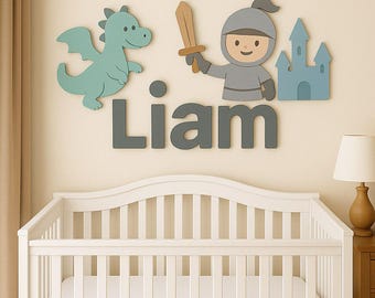 May include: A nursery wall decoration featuring a dragon, knight, and castle in shades of blue and gray. The name "Liam" is spelled out in gray letters. A white crib and lamp are in the foreground.
