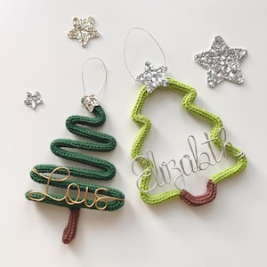 May include: Two handmade Christmas tree decorations. One is a green knitted tree with a gold wire "Love" inscription. The other is a green knitted tree outline with the name "Elizabeth" in silver wire. Glittery star accents.