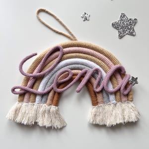 May include: A wall hanging featuring a rainbow made of coiled rope in shades of tan, pink, white, and grey, with a fringe at the bottom. The name Lena is written in cursive using pink rope across the centre, accented by two silver glitter stars.
