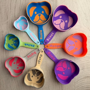 Puede incluir: Una colección de cucharas con forma de perro en varios colores, incluyendo azul, verde, morado, naranja y gris. Cada cuchara presenta un diseño de cara de perro y un nombre en el mango. Las cucharas están dispuestas en un patrón circular.