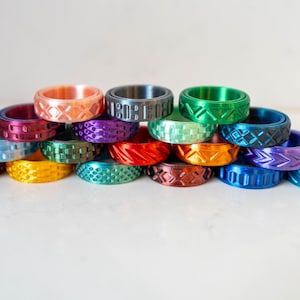 May include: A collection of metal rings in various vibrant colors including blue, green, orange, red, purple, and yellow. Each ring features a distinct geometric pattern engraved into the surface, such as chevrons, cross-hatching, and dashes.