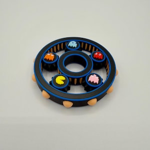 Könnte beinhalten: Ein schwarzer, blauer und beigefarbener Fidget Spinner mit bunten Pac-Man-Geistern. Der Spinner hat ein kreisförmiges Design mit einer zentralen Nabe und sechs kleineren Pac-Man-Geisterfiguren in Orange, Blau, Rot, Rosa und Gelb.