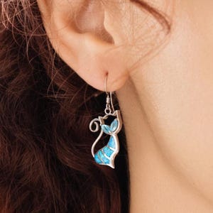 Pode incluir: Brincos prateados em forma de gato com incrustações azuis e turquesas. Os brincos apresentam uma silhueta de gato estilizada com uma cauda curva e um gancho pendurado. A incrustação azul é visível no corpo e na cabeça do gato.
