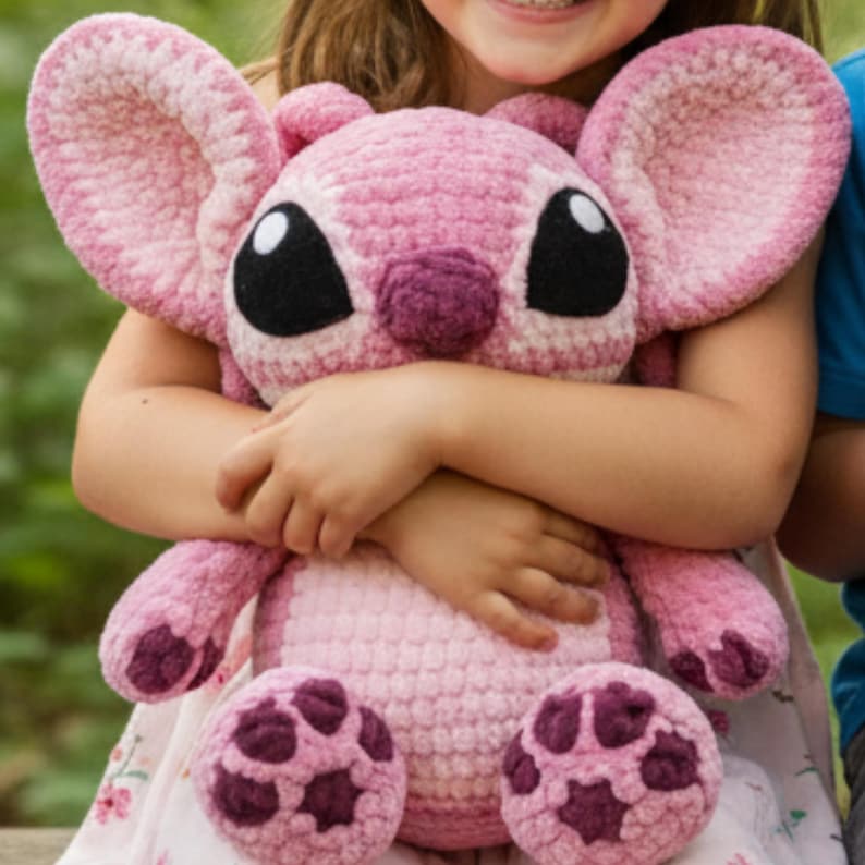 May include: A large, pink crocheted plush toy with large ears, black eyes, and a purple nose. The toy has a round body and is being held by a person. The plush toy's feet have a dark purple pattern.