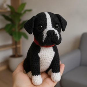 May include: A small, handcrafted crochet dog figurine featuring black and white yarn. The dog has a white stripe on its face, a white chest, and white paws. It wears a solid red collar around its neck and has two large, round, black plastic eyes.