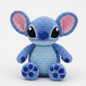 May include: A handmade blue crochet plush toy with large black eyes, pink-lined ears, and a small nose. The plush has a light blue belly and darker blue feet. The toy is sitting on a white surface.