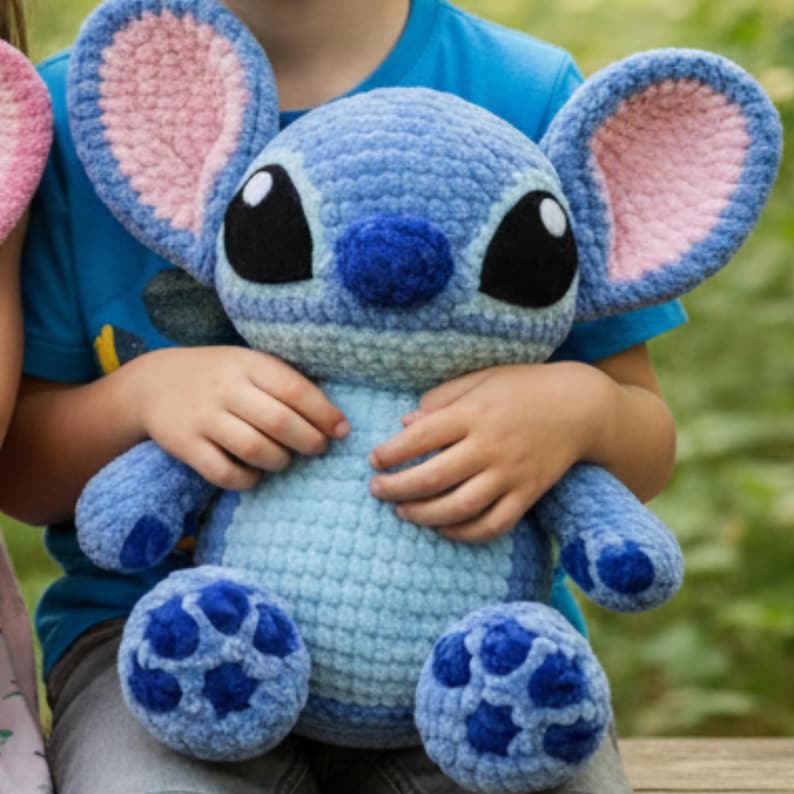 May include: A handmade blue crochet plush toy with large ears, black eyes, and a dark blue nose. The plush has light blue and dark blue accents. The toy is being held by a person wearing a blue shirt. The toy is a character from a popular animated film.