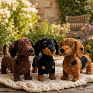 Op de afbeelding: Drie gehaakte teckel hondjes in bruin, zwart en beige. De honden hebben slappe oren en kleine staarten en staan op een crèmekleurig, getextureerd oppervlak. De achtergrond toont groen en een stenen muur.
