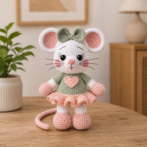 Puede incluir: Una muñeca de ratón tejida a ganchillo con pelaje blanco, orejas grandes con centros rosas y un lazo verde salvia. Lleva una camisa verde salvia con un corazón rosa aplicado y una falda rosa con volantes. El ratón tiene ojos negros y una cola larga.