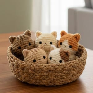 May include: A woven basket filled with five crocheted cat toys. The cats are various colors, including brown, cream, and orange, with black eyes and small ears. The basket is a light brown color and sits on a wooden surface.