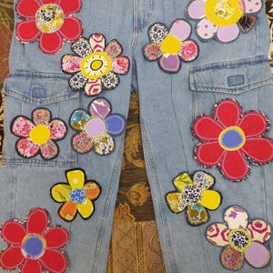 May include: Light blue denim cargo pants adorned with colorful, appliqued flower designs. The flowers feature various patterns and colors, including red, yellow, purple, and floral prints, creating a unique and artistic look. The pants have pockets and a classic denim style.