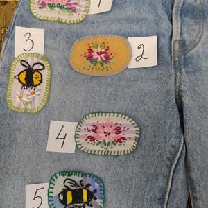 May include: Light blue denim jeans with five embroidered patches. The patches feature floral and bee designs in various colors, including pink, yellow, and black. The patches are oval-shaped and sewn onto the jeans.