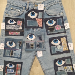 May include: Light blue denim jeans featuring eight distinct rectangular fabric patches numbered one through eight. Each patch displays a hand-stitched blue eye motif surrounded by various fabric scraps in different textures, patterns, and colours.