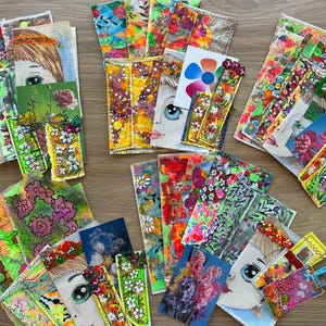 以下が含まれることがあります： 大きな目をした手描きの顔、鮮やかな花柄、抽象的な色の飛沫、花の写真プリントなど、多様な芸術的デザインが施された長方形のしおりのコレクション。それらが散らばった状態で配置されています。