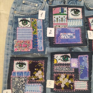 Pode incluir: Calças de ganga azul claro com vários patches de tecido costurados na frente. Cada patch apresenta um design único, incluindo um olho, padrões florais e formas geométricas. Os patches são em várias cores, incluindo rosa, azul e branco.