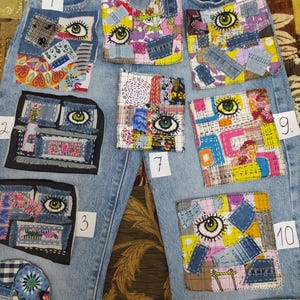 May include: Light blue denim jeans with eleven patches of various sizes and designs. Each patch features an embroidered eye with green iris and black outline. The patches are made of fabric scraps and denim, with colorful patterns and textures.