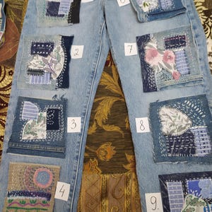 May include: Light blue denim jeans adorned with nine decorative patches. Each patch showcases a unique design with floral and geometric patterns, using diverse fabrics and stitching methods. The patches feature shades of blue, white, pink, and purple.