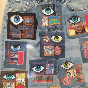 May include: Light blue denim jeans featuring multiple rectangular fabric patches sewn onto the front. The patches display various eye illustrations in blue and green, combined with colorful floral, plaid, and patterned fabric scraps with visible stitching.