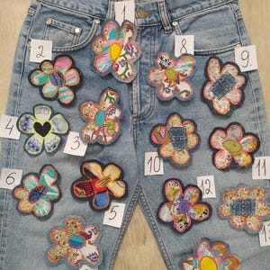 May include: Light blue denim jeans featuring fourteen distinct, colorful, fabric flower patches arranged across the front. Each patch is numbered with a small white tag, showcasing various patterns, textures, and stitched edges in a variety of colors.