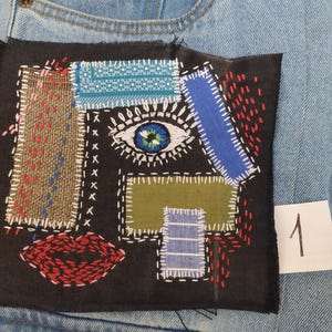 May include: Close-up of a denim jean pocket with a black patch featuring an embroidered abstract face. The patch includes a blue eye, red lips, and various colorful fabric patches. A white number 1 is visible.