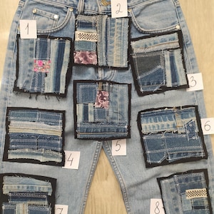 May include: Light blue denim jeans featuring eight distinct rectangular patchwork squares sewn onto the front. Each patch is composed of various blue denim fabric scraps, black fabric borders, and small numbered white paper labels from 1 to 8.